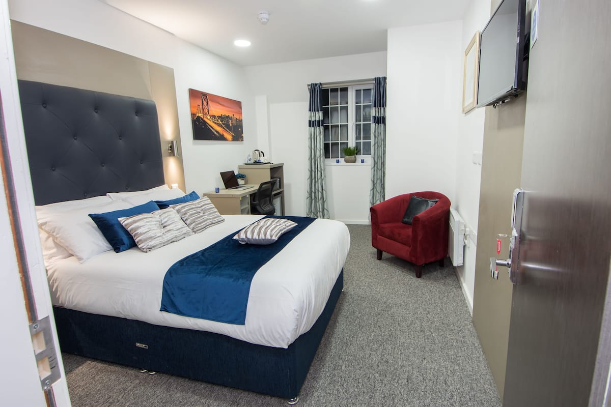 Ein Hotelzimmer mit einem großen Bett, einem Schreibtisch, einem Sessel, einem Fernseher und einem Fenster mit Vorhängen.