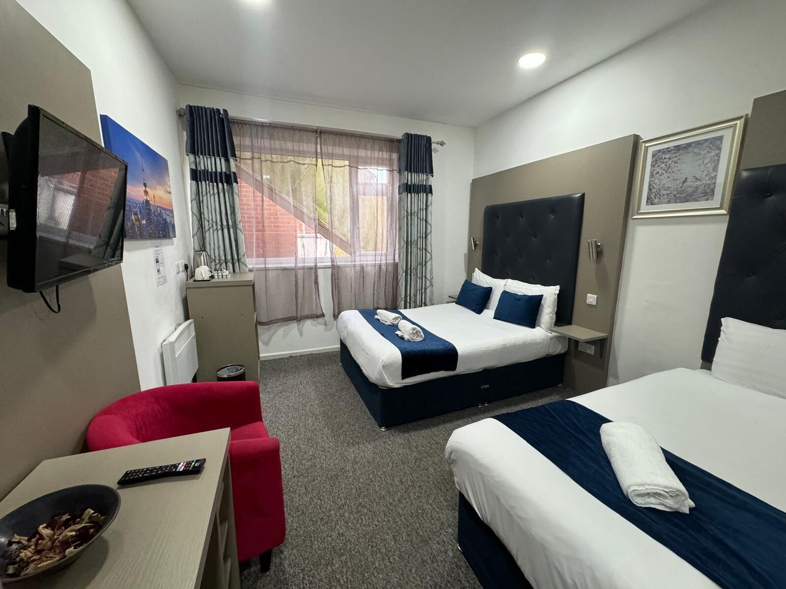A hotel room with two beds, a TV, a desk with a bowl of fruit, and a window with curtains.