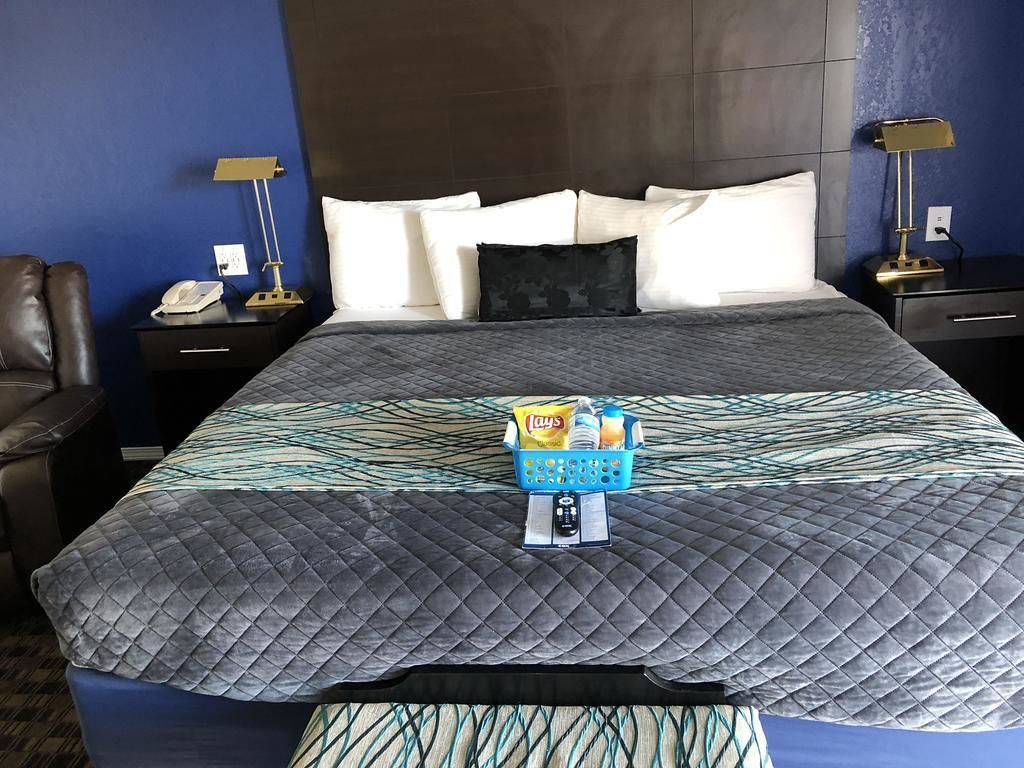 A hotel room with a large bed, a nightstand with a lamp, a chair, and a small table with a phone and a basket of snacks.