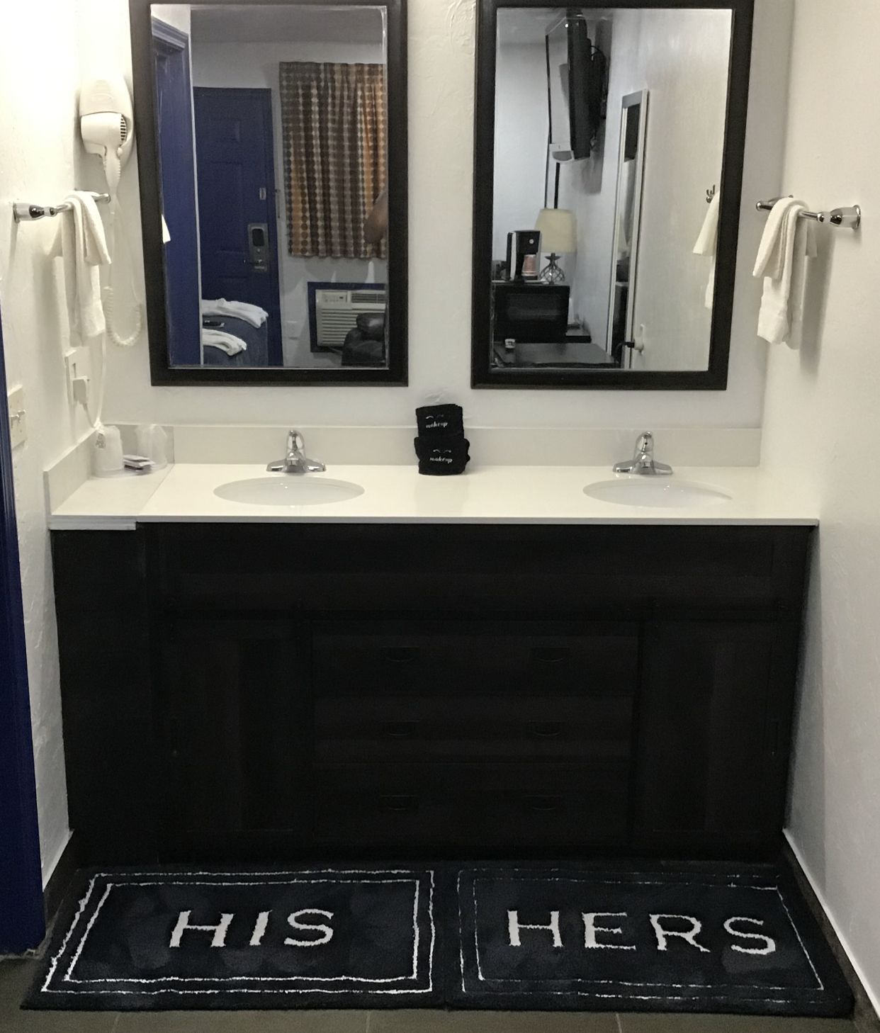 Un baño con dos lavamanos, un secador de pelo y un espejo. Hay una alfombra negra con la inscripción 'HIS HERS'.