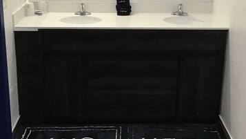 Un baño con dos lavamanos, un secador de pelo y un espejo. Hay una alfombra negra con la inscripción 'HIS HERS'.