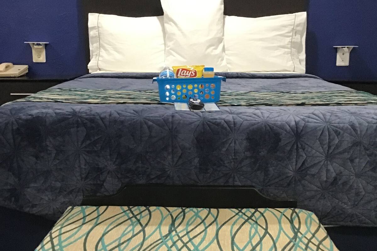 A hotel room with a bed, pillows, a blue basket with snacks, and a patterned bench.