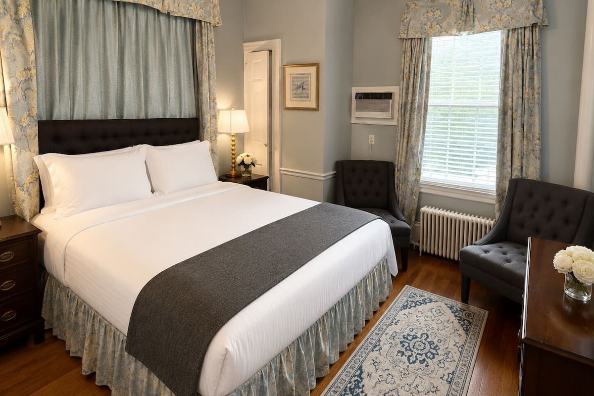 A neatly arranged bedroom with a large bed, a chair, a nightstand, a lamp, a radiator, and a window with curtains.