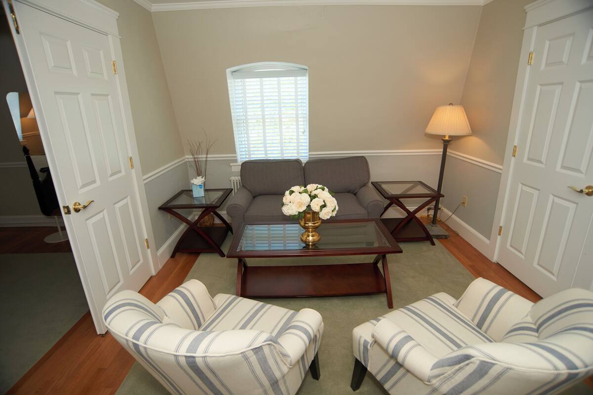 A living room with a sofa, two armchairs, a coffee table with a vase of flowers, a lamp, and a window with blinds.