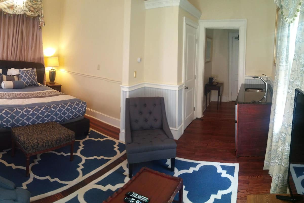 A hotel room with a large bed, a grey armchair, a small table, and a desk with a sink.