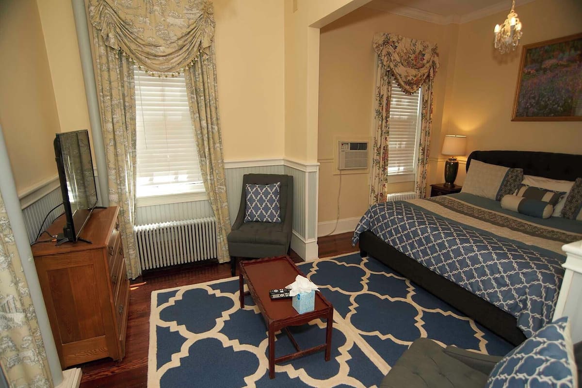 A hotel room with a large bed, a sofa, a small table, and a chair. There are two windows with curtains, a radiator, and a painting on the wall.