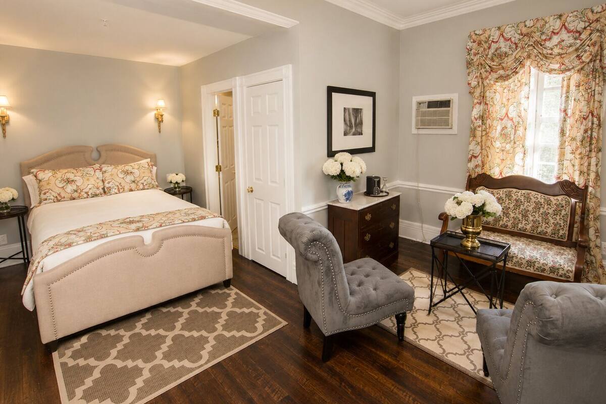 A bedroom with a bed, two armchairs, a dresser, and a desk with a lamp.