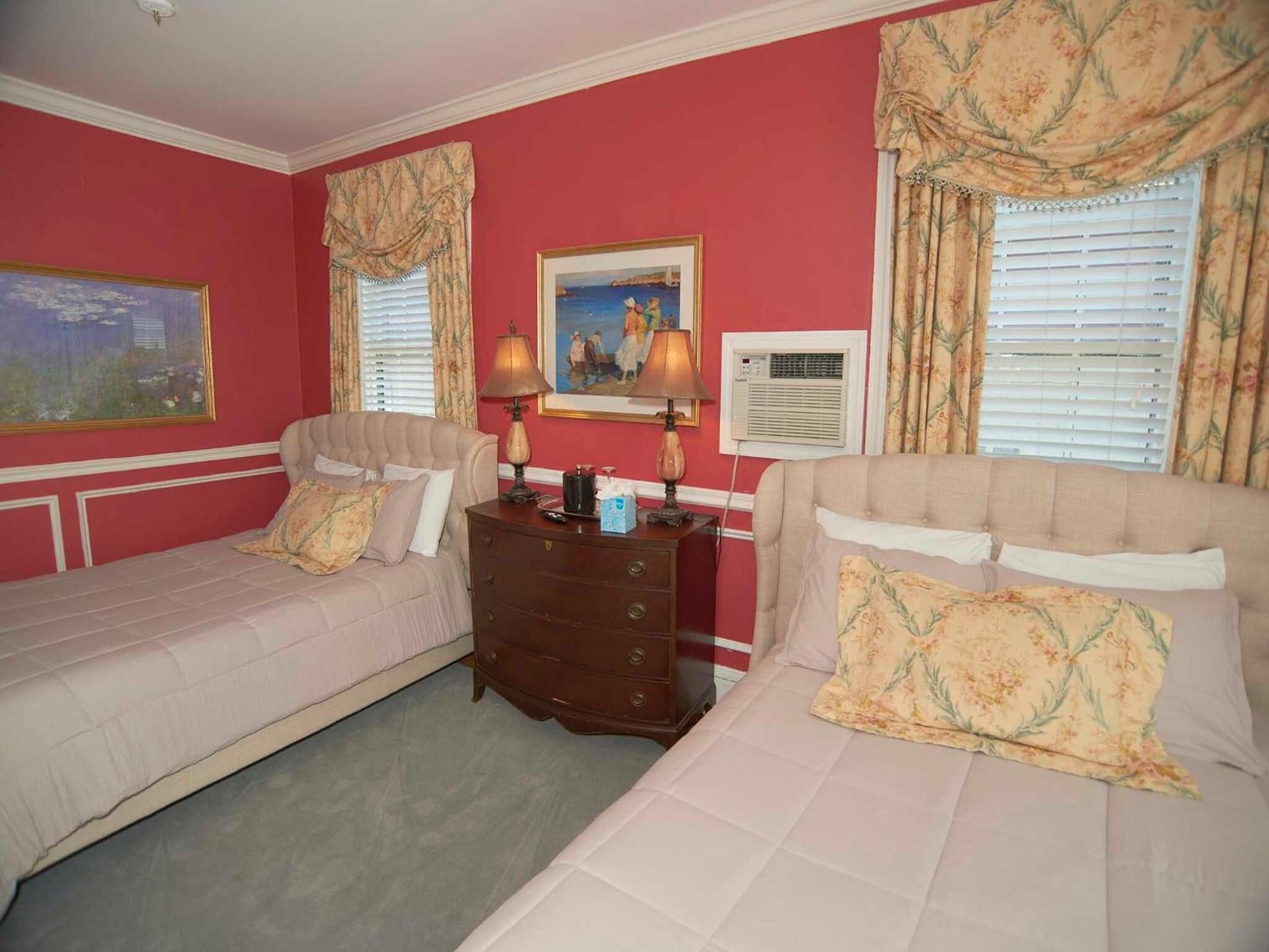 A hotel room with two single beds, a wooden dresser, a painting, a lamp, and an air conditioning unit.