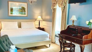 A hotel room with a bed, a desk, a chair, a chandelier, a television, and a painting on the wall. - Bellevue Manor (Newport)