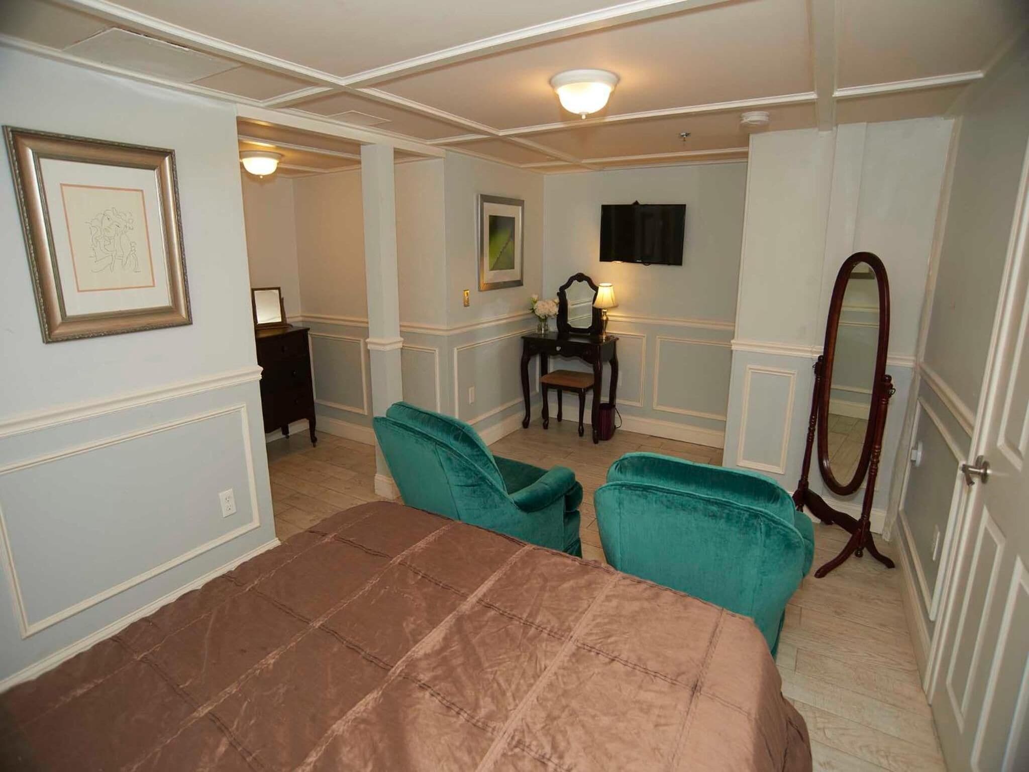 A bedroom with a large bed, two teal armchairs, a wooden dresser, a vanity with a mirror, and a television mounted on the wall.