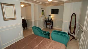 A bedroom with a large bed, two teal armchairs, a wooden dresser, a vanity with a mirror, and a television mounted on the wall.