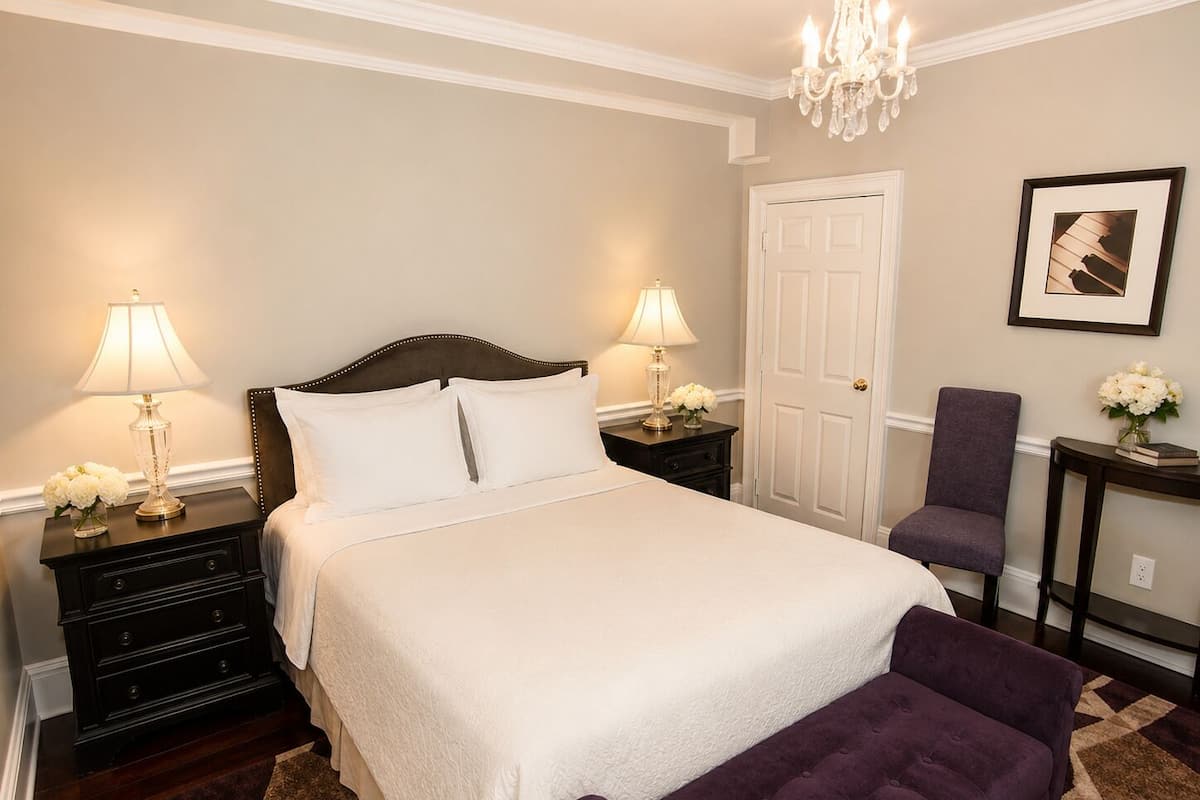 A neatly arranged bedroom with a bed, two bedside tables with lamps, a chair, a door, and a framed picture on the wall.