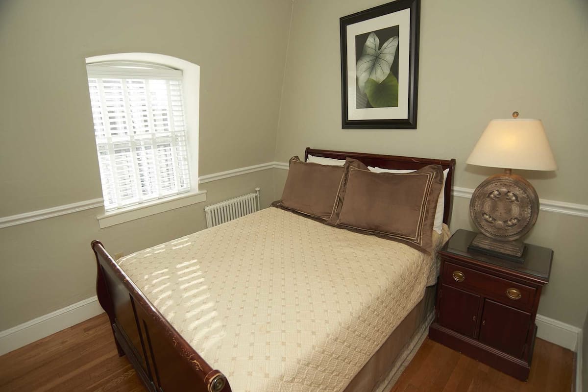 A bedroom with a wooden bed, a nightstand with a lamp, a window with blinds, and a framed picture on the wall.