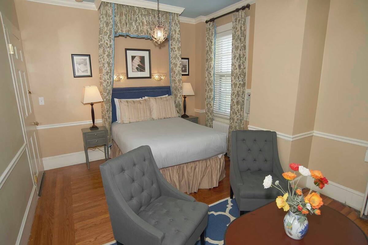 A hotel room with a bed, two armchairs, a small table with a vase of flowers, and a chandelier.