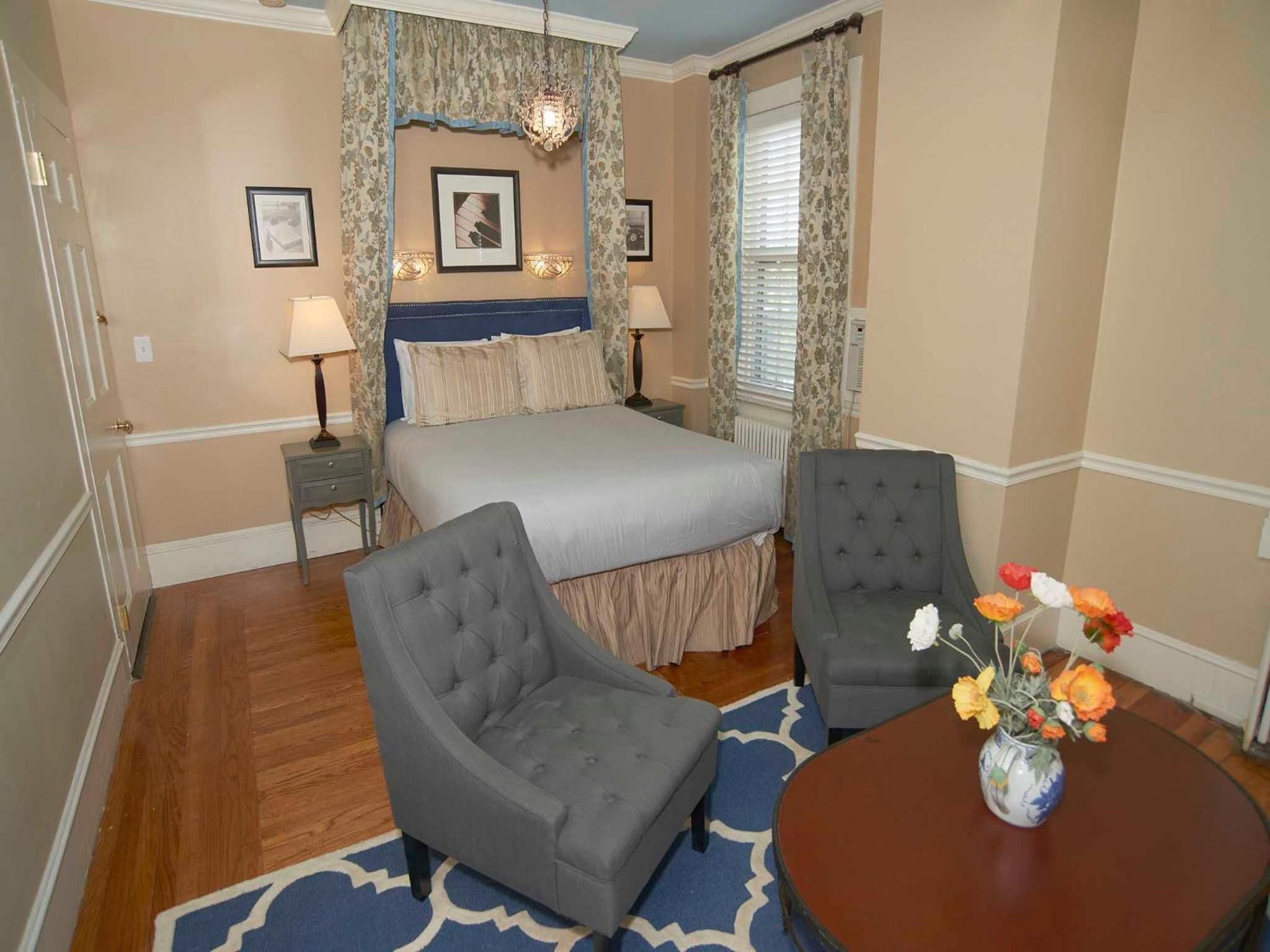 Un dormitorio con cama, dos sillones, una mesita de noche, lámpara, un florero con flores y un candelabro.