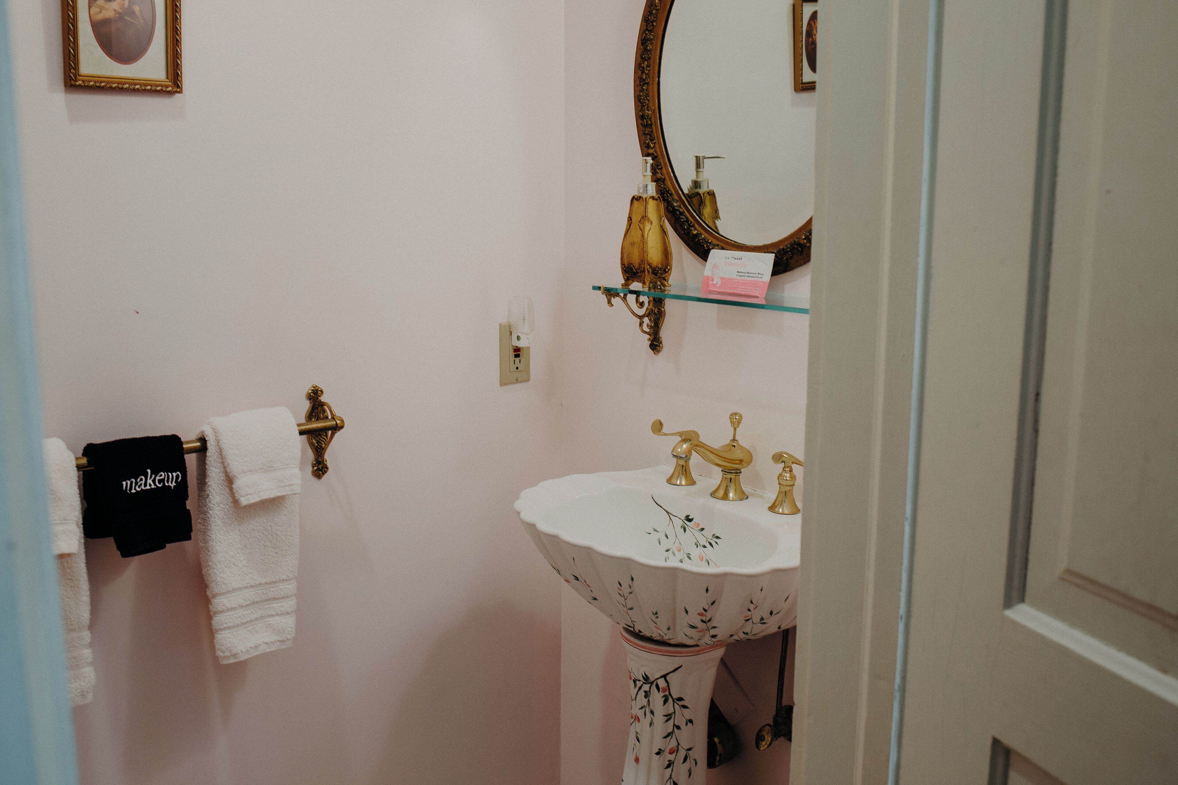 A bathroom with a white sink, gold faucets, and a mirror with a gold frame. There is a towel rack with white and black towels, and a small shelf with a glass container and a pink box.