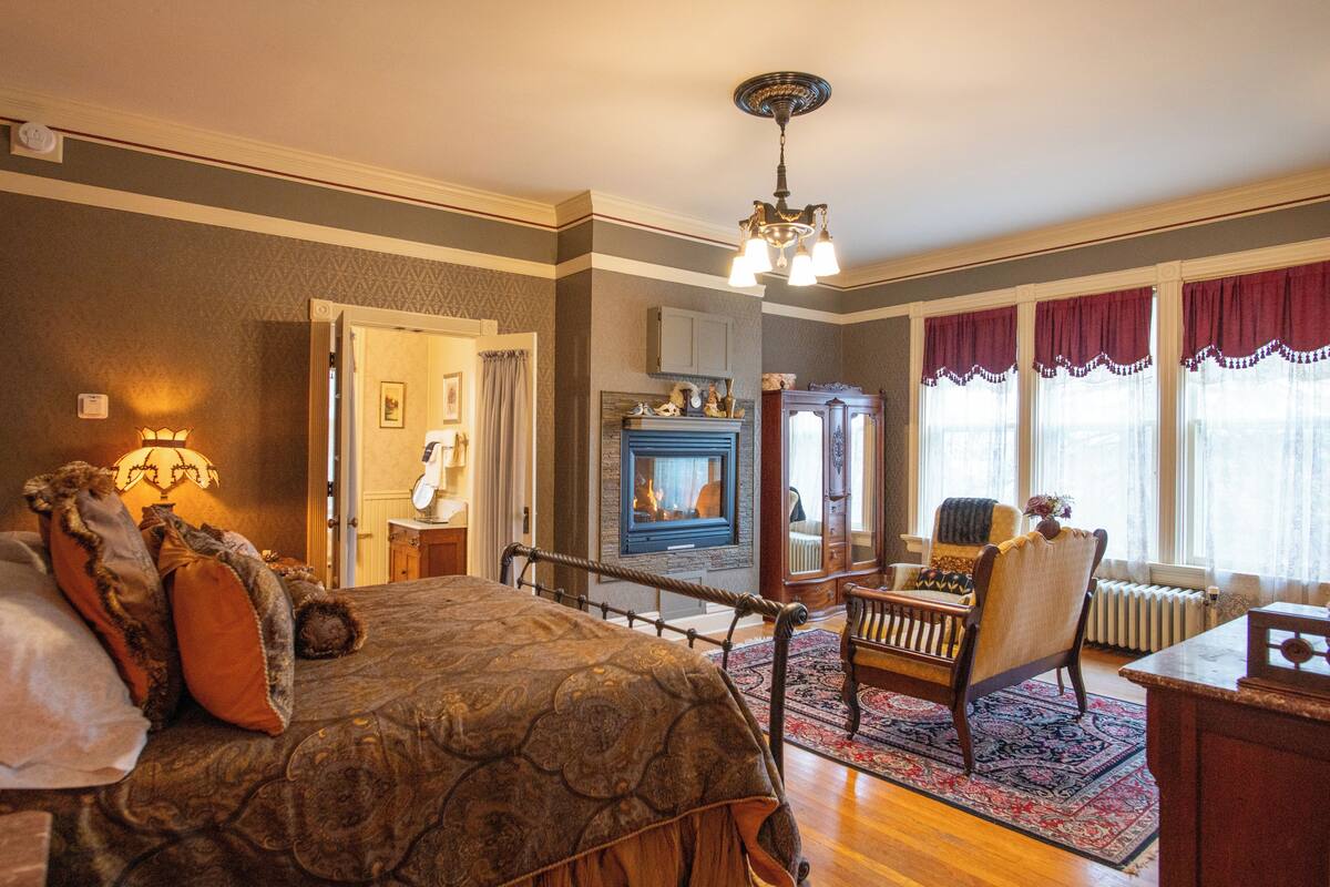 Une chambre à l’ancienne, dotée d’une cheminée, d’un lit avec une literie à motifs, d’une chaise en bois et offrant une vue sur l’extérieur grâce à de grandes fenêtres.