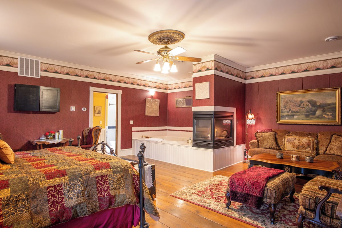 Une pièce comprenant un lit, un canapé, une cheminée, un ventilateur de plafond et un tableau au mur.