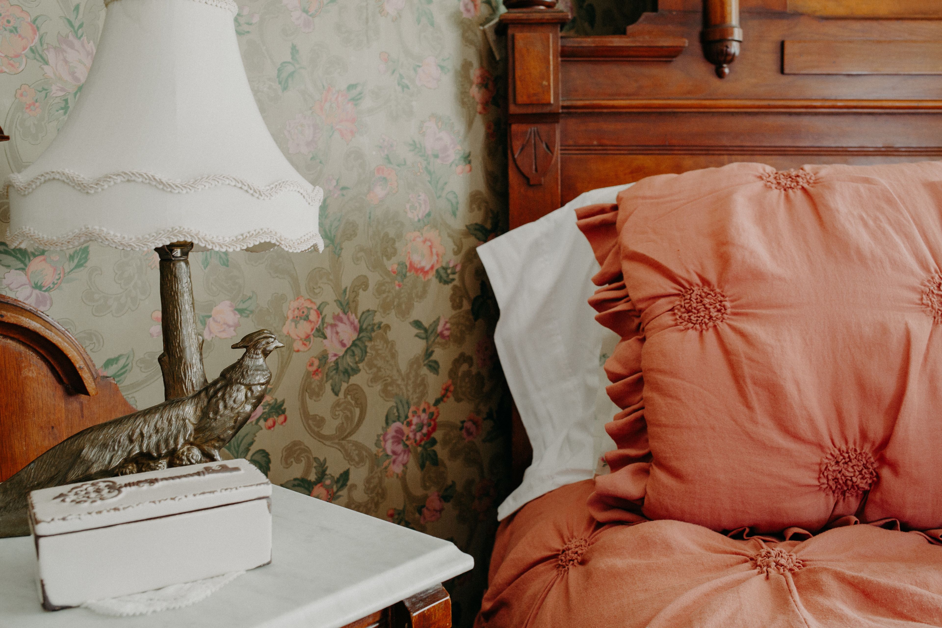 Une chambre avec un papier peint à motifs floraux, une commode en bois, une lampe et un coussin moelleux de couleur rose.