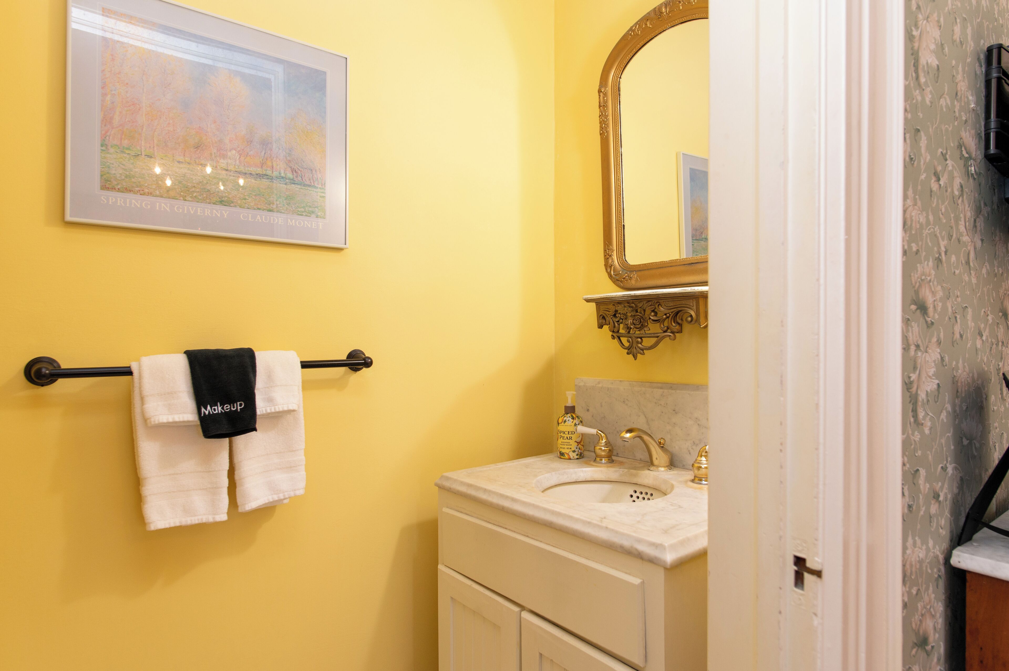 Un baño con pared amarilla, un cuadro enmarcado, un perchero con toallas blancas, un lavamanos con grifo dorado y un espejo con marco ornamentado.