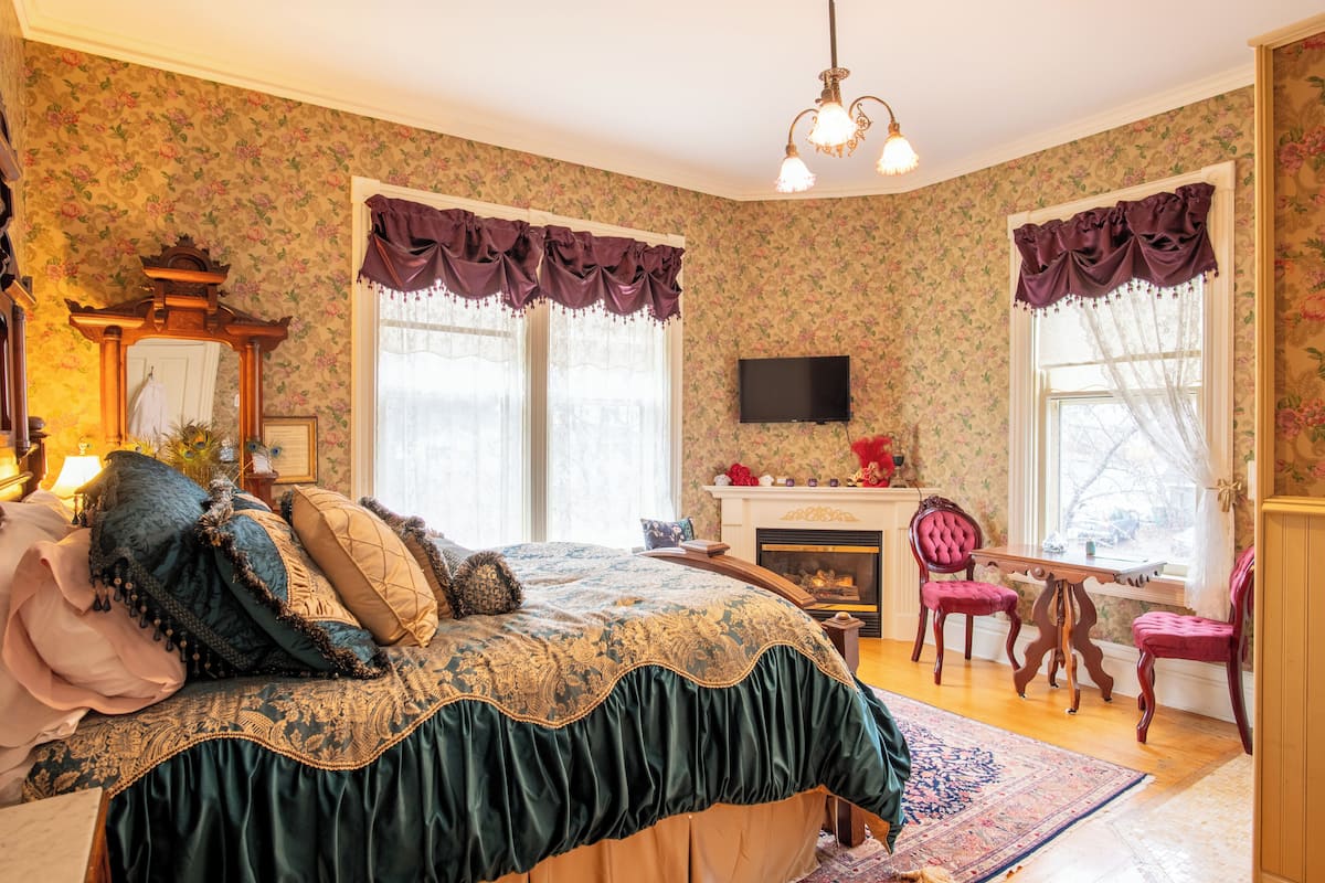 Une chambre à coucher avec un grand lit, une cheminée, deux fauteuils et un téléviseur fixé au mur.