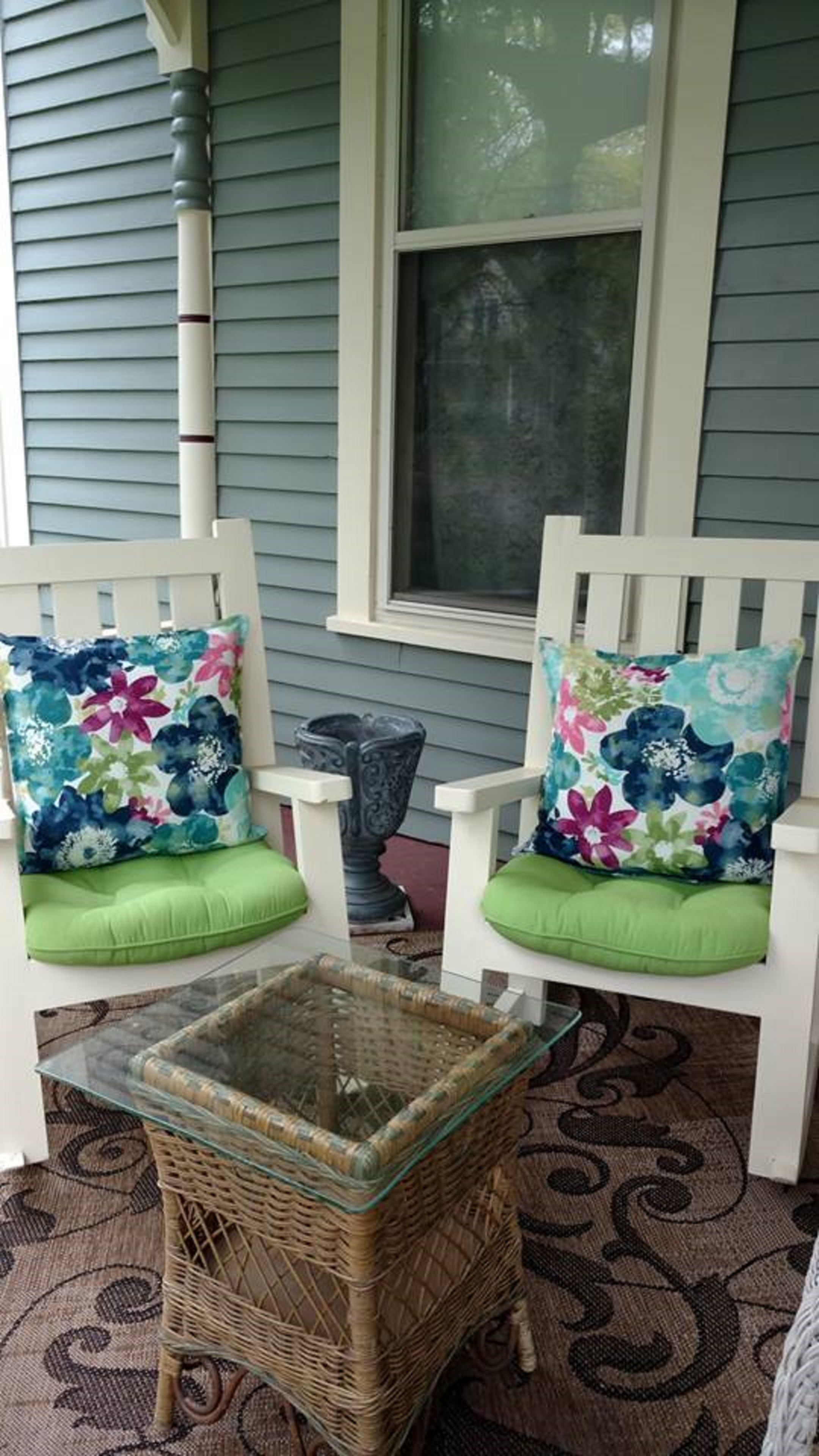 Two white porch chairs with floral cushions and green seat cushions, a wicker table, and a window with a view of trees.