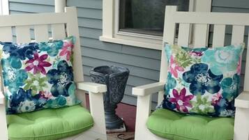 Two white porch chairs with floral cushions and green seat cushions, a wicker table, and a window with a view of trees.