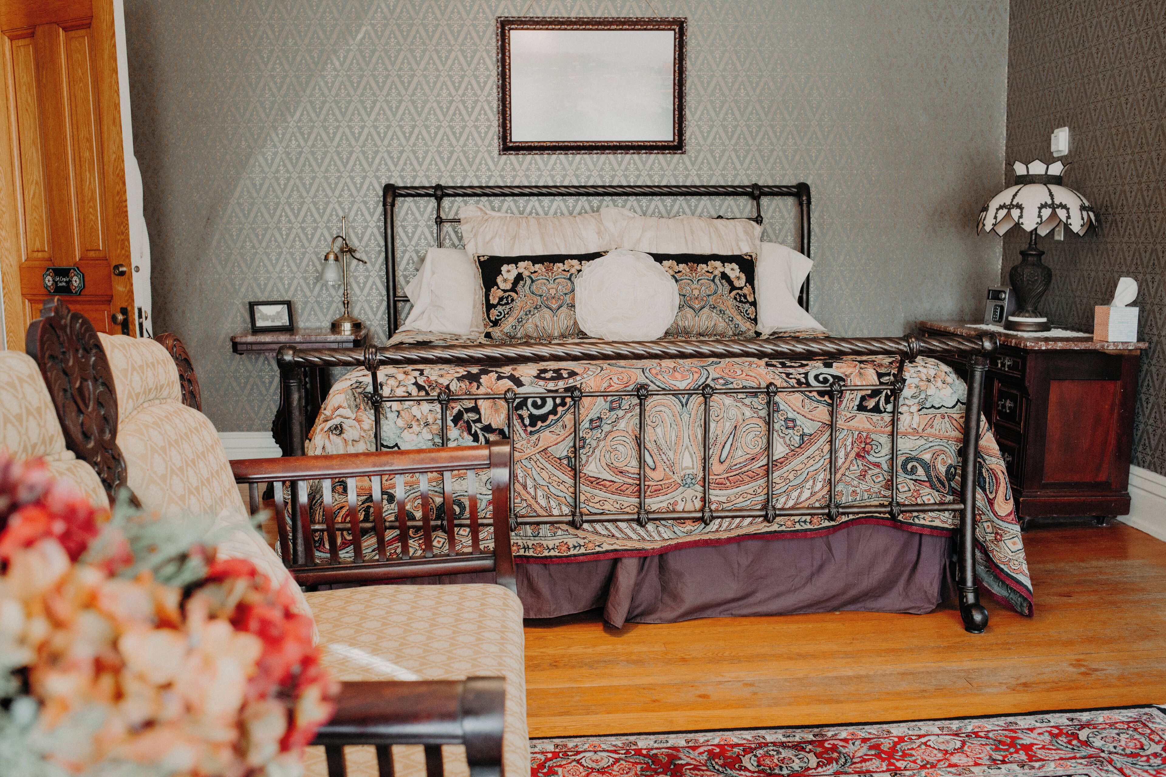 A bedroom with a four-poster bed, a wooden chair, a nightstand with a lamp, and a patterned rug.