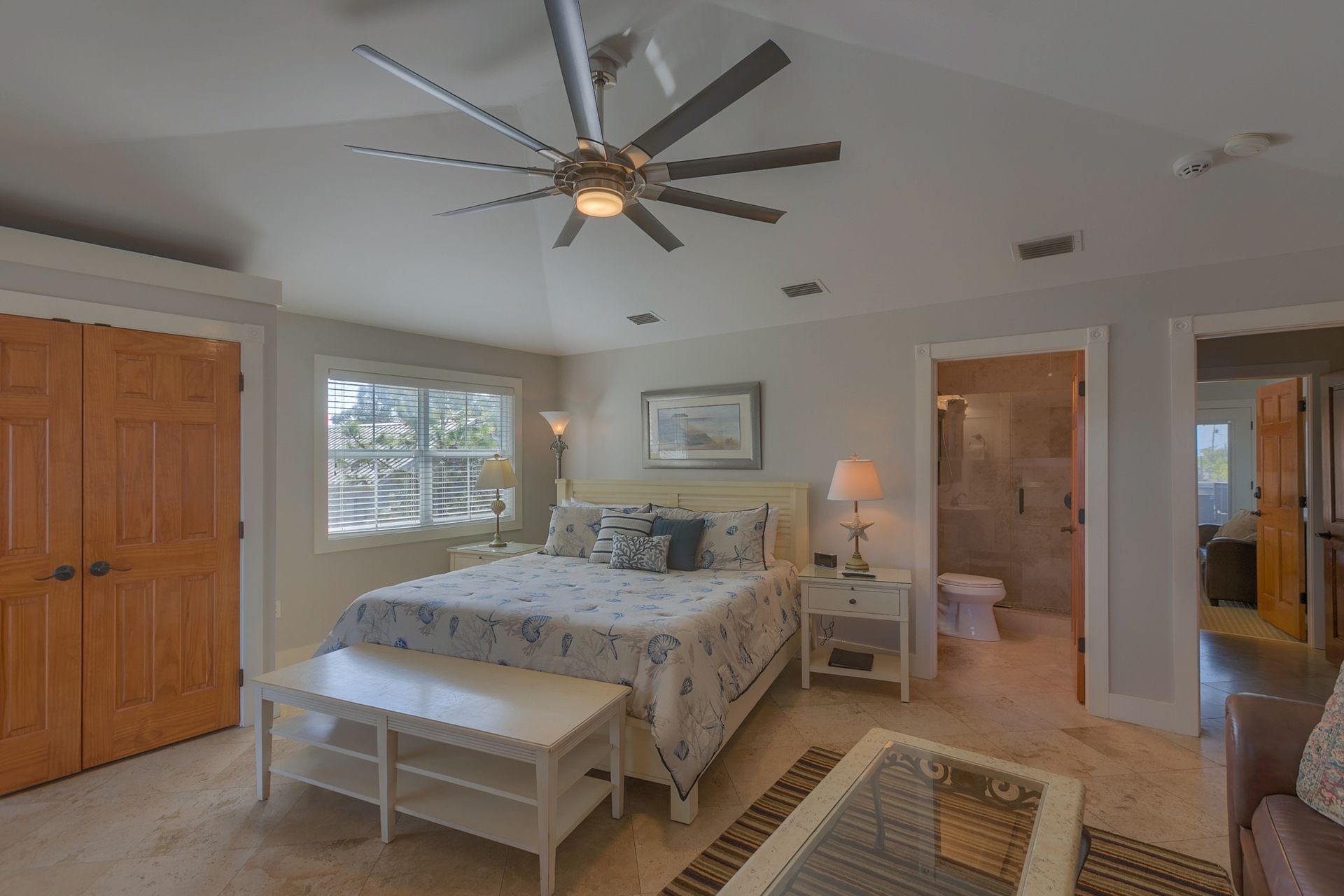 A bedroom with a bed, bedside tables, a desk, a ceiling fan, a window, and a bathroom with a toilet and shower.