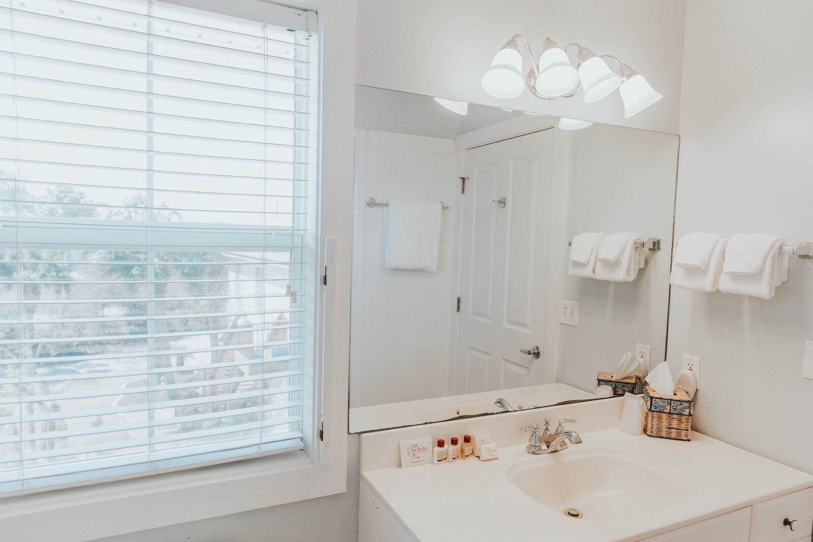 Un baño con un espejo grande, lavamanos blanco y una ventana con persianas.