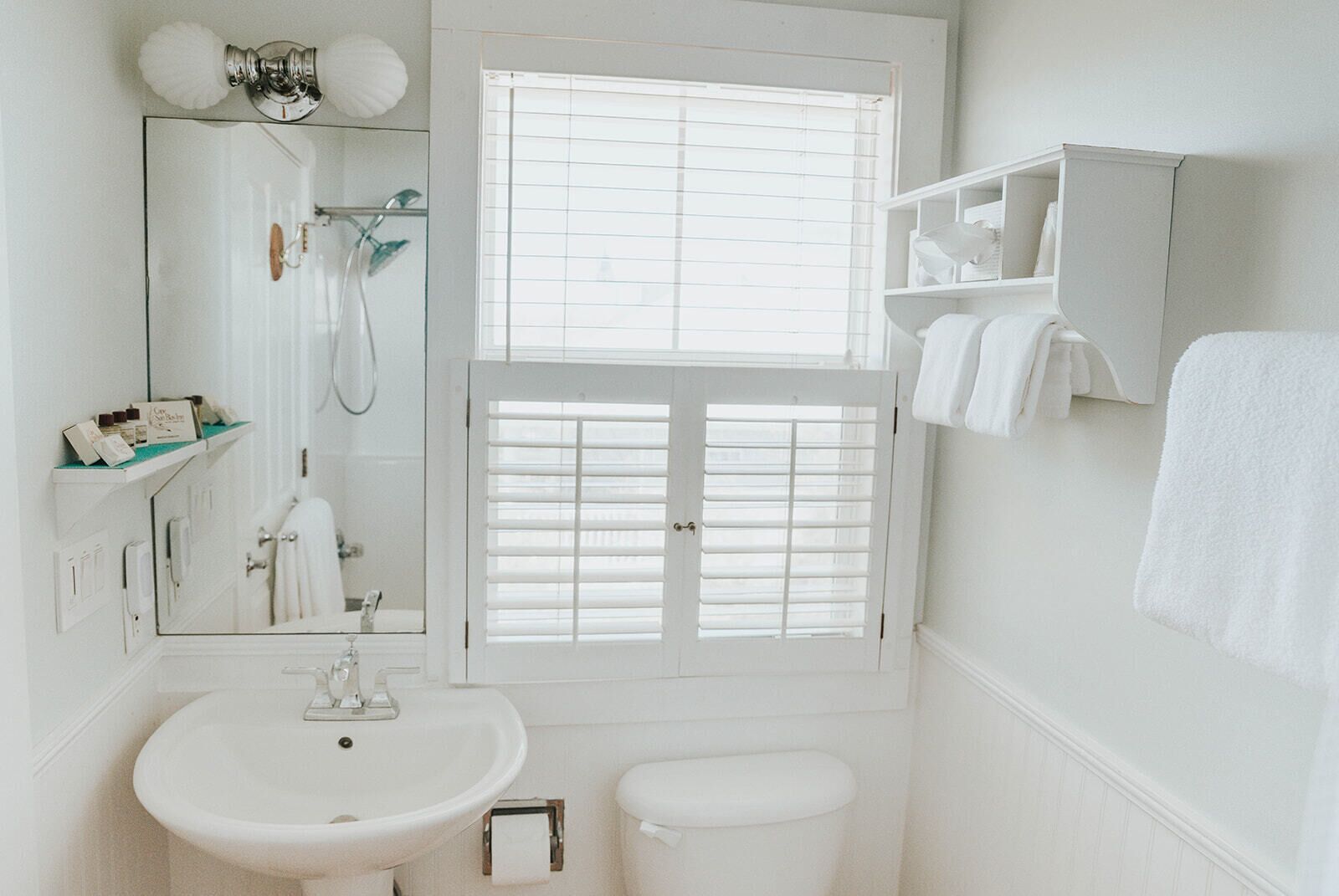 Un baño blanco con lavamanos, inodoro y ventana con persianas.