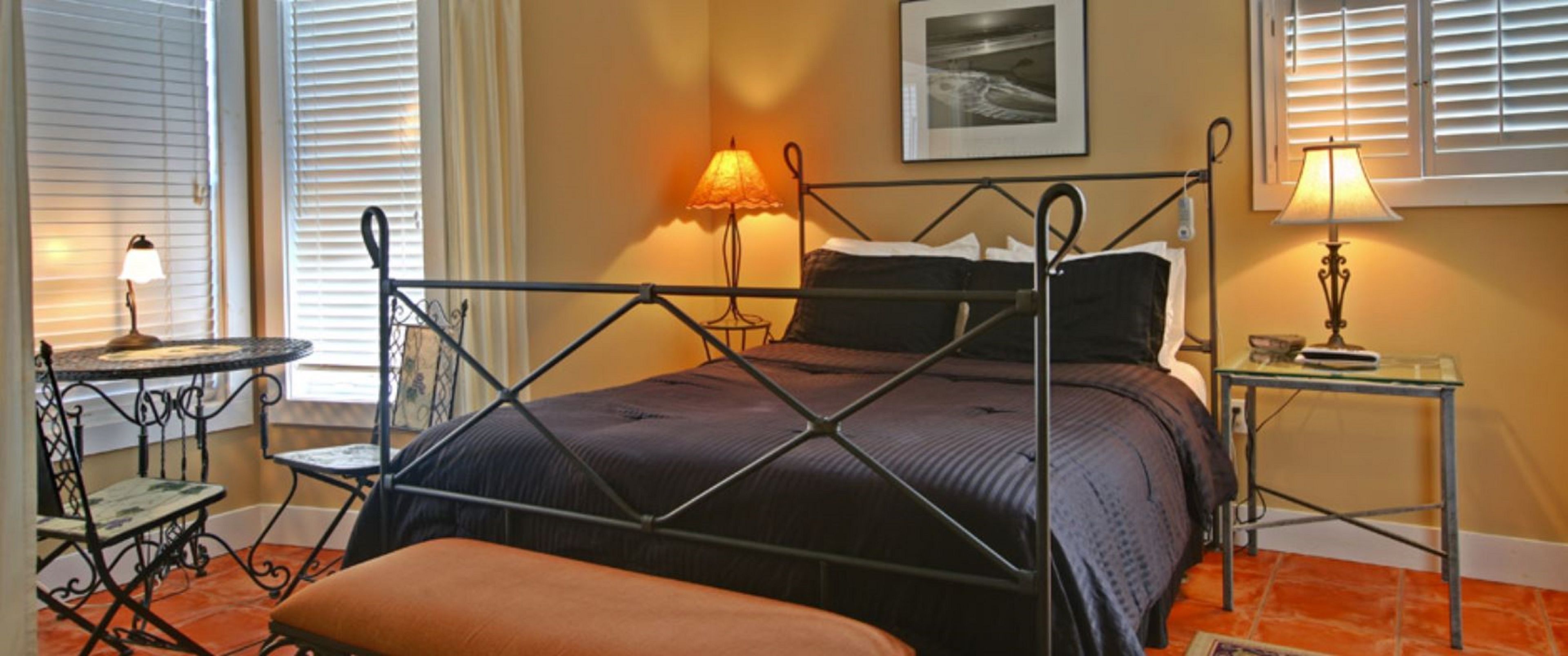 A neatly arranged bedroom with a four-poster bed, a small round table with a chair, and a bedside table with a lamp.