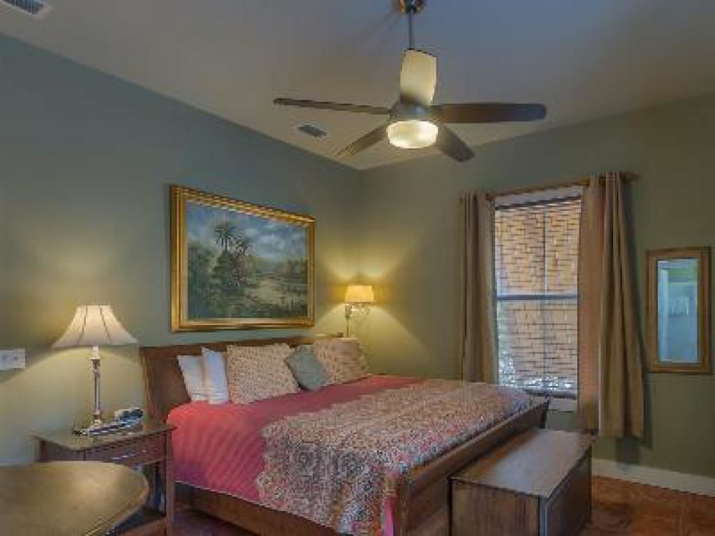 A bedroom with a bed, a nightstand, a lamp, a ceiling fan, a window with curtains, and a painting on the wall.