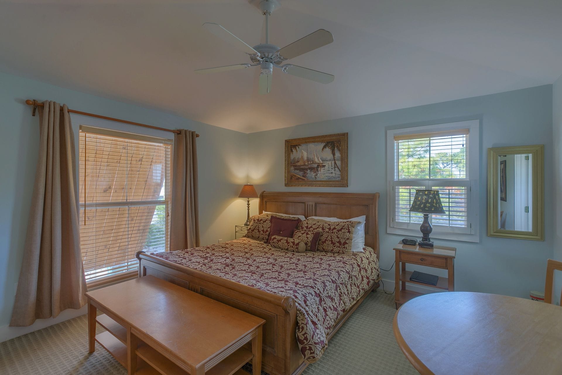 A bedroom with a bed, a nightstand, a table, a ceiling fan, and a painting on the wall.