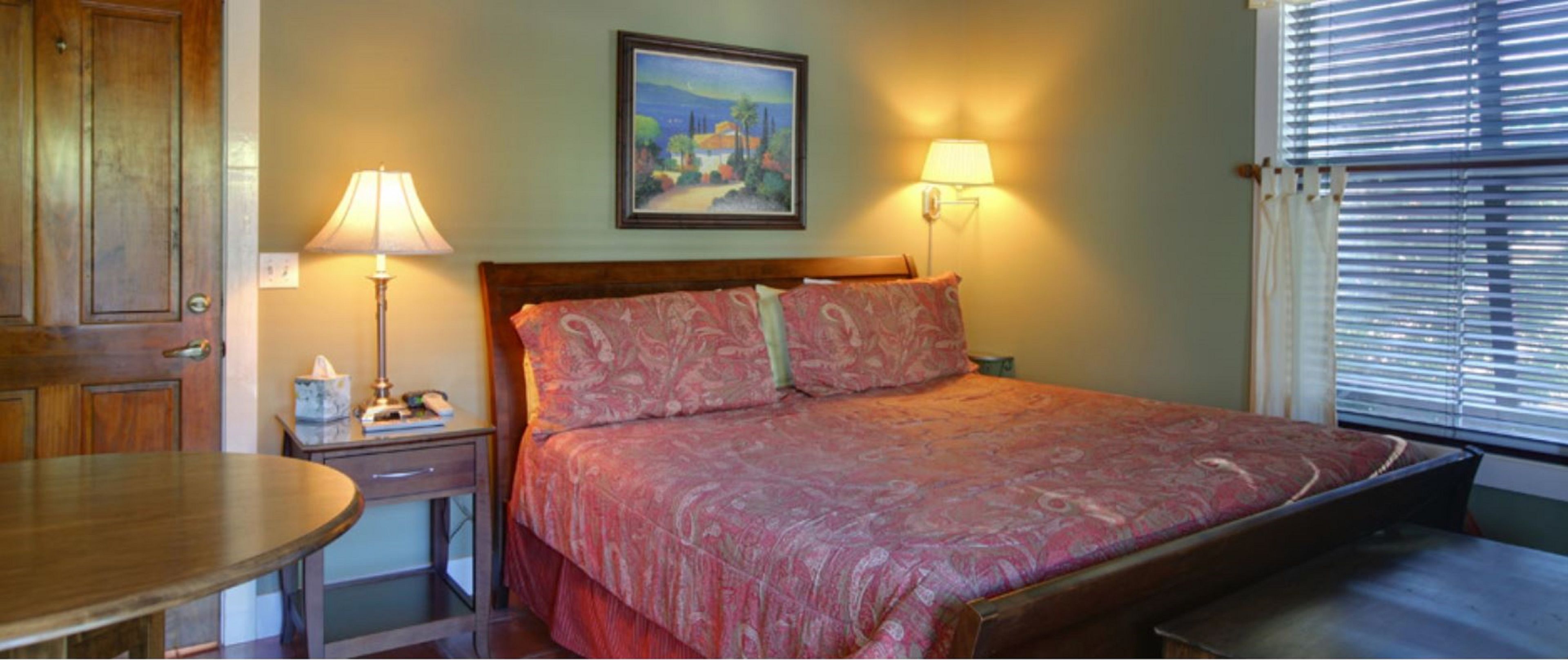 A bedroom with a bed, bedside lamps, a painting on the wall, and a window with blinds.