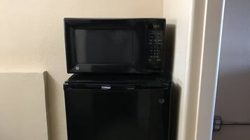 A black refrigerator with a microwave on top, located in a corner of a room with tiled flooring and a door leading to another area.