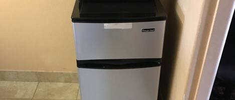 A compact refrigerator with a microwave on top, situated in a corner against a wall with a tiled floor.