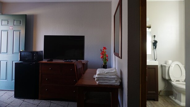 A hotel room with a television, a small refrigerator, a wooden dresser, a stack of towels, a potted plant, and a bathroom visible through an open door.