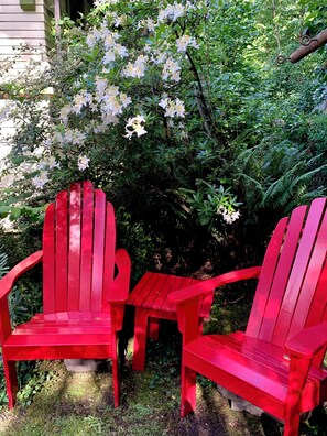 Zwei rote Adirondack-Stühle, ein kleiner roter Tisch und ein üppiger, grüner Busch mit weißen Blüten.