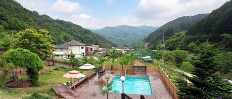 Una piscina rodeada por una terraza de madera, con vistas a un paisaje montañoso y casas al fondo.