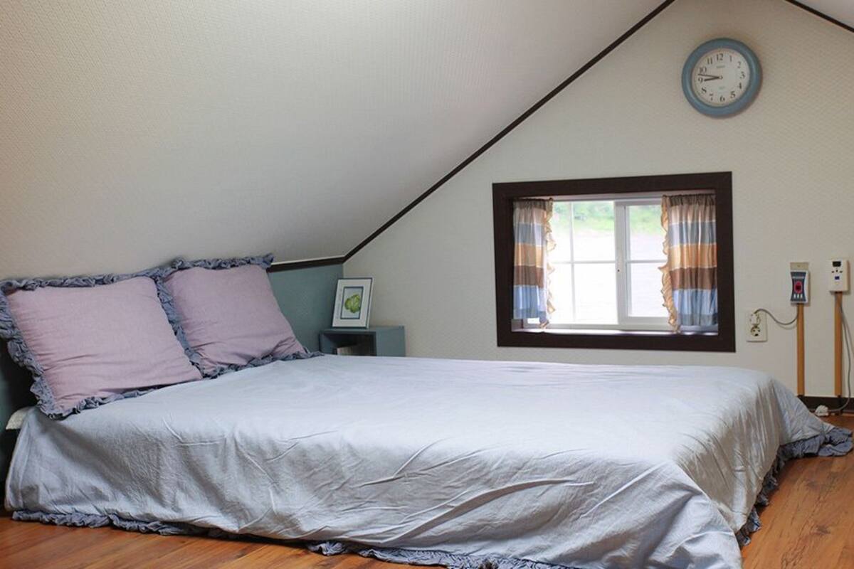 Un dormitorio con cama, una ventana con cortinas, un reloj en la pared y una mesita de noche con un marco de fotos.
