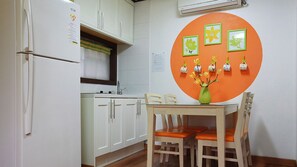 Une cuisine avec des meubles blancs, un réfrigérateur, une table à manger avec des chaises orange et un mur orné de tableaux.