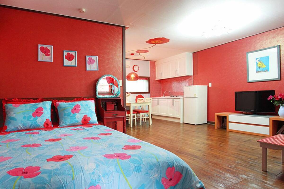 Un dormitorio con cama, escritorio, silla, televisor y una zona de cocina.