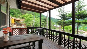 A wooden deck with a table and chairs, a grill, and a view of a lush green landscape with trees and a house in the background. - Duo Pension (Pyeongchang)