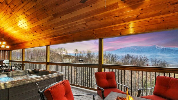 A wooden deck with a stone fire pit and red chairs, overlooking a mountainous landscape.