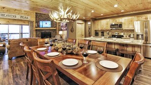 A spacious dining area with a wooden table set for six, surrounded by wooden chairs. A chandelier hangs above the table, and a television is mounted on the wall.