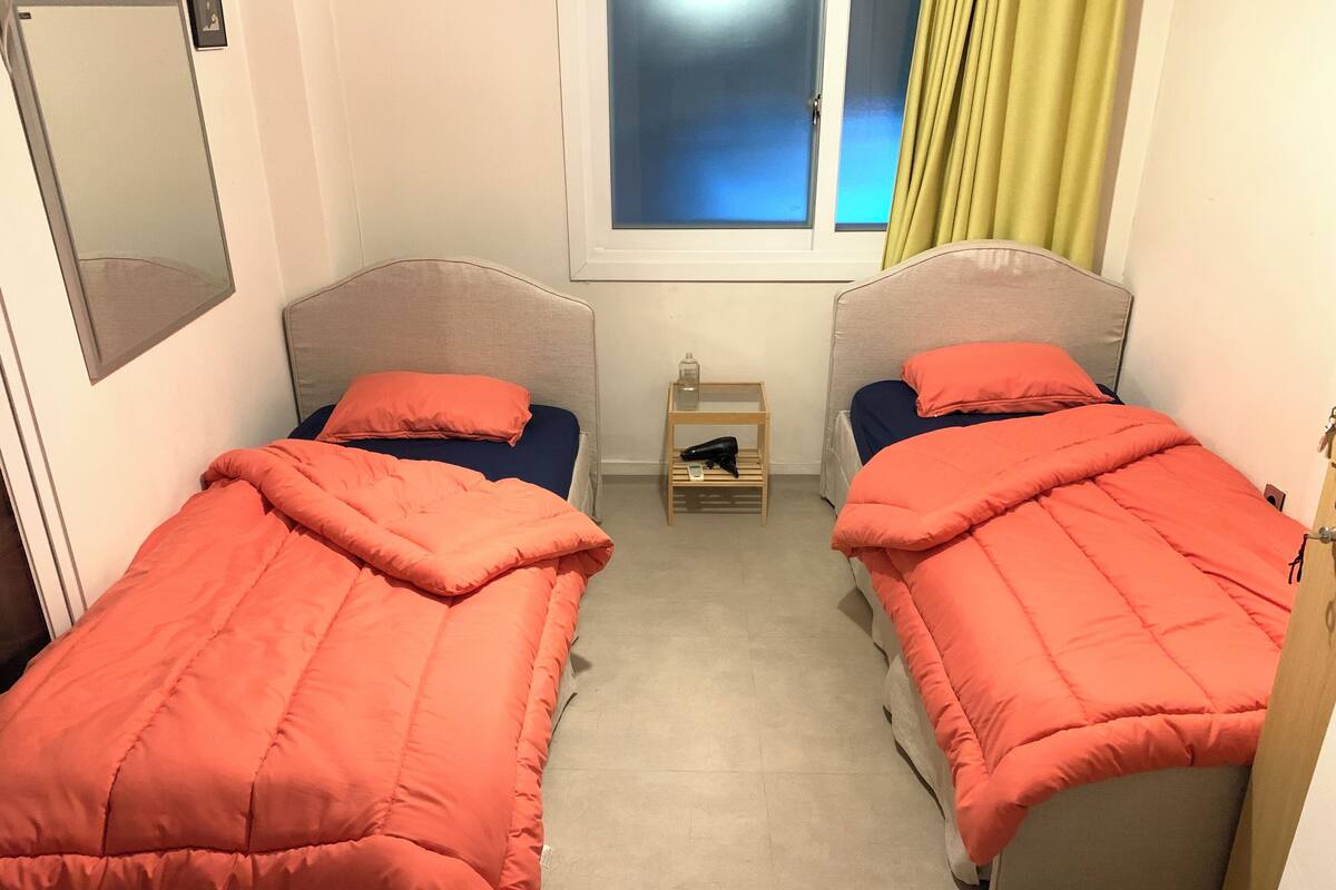 A small, well-lit room with two single beds, each with a matching orange blanket and pillow. There is a small wooden nightstand with a lamp between the beds, and a window with a blue view and yellow curtains.