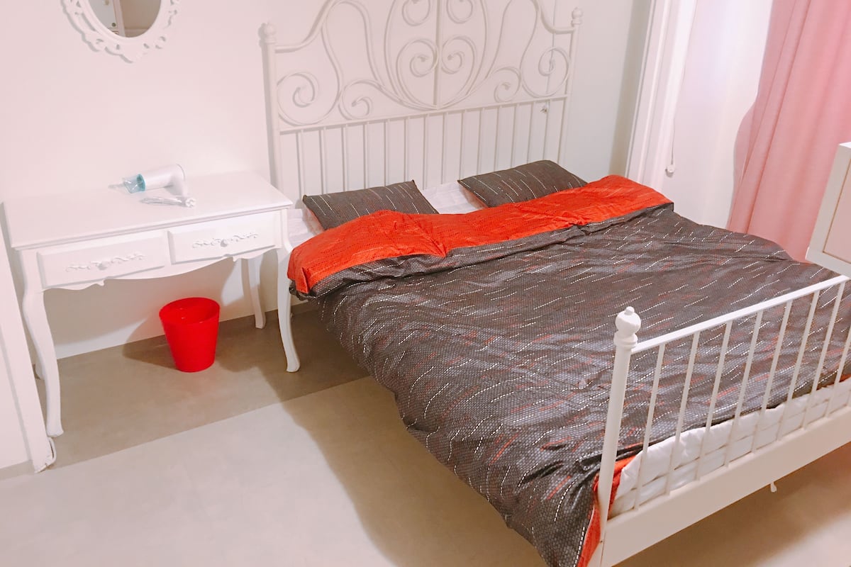 A bedroom with a white metal bed frame, a red bucket, a white desk with drawers, and a mirror on the wall.