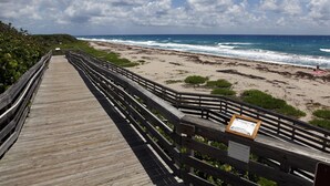 A wooden boardwalk leading to a beach with a wooden railing and a sign. - Restoration Hardware PGA National Condo w/ 180° Golf Course Views (Palm Beach Gardens)
