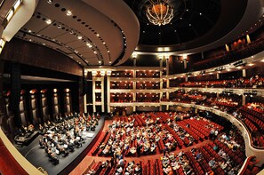 Een ruime binnenzaal met terrasvormige zitplaatsen, een podium en een grote kroonluchter.